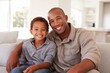 © Supanat - Happy African American father and son smiling together sitting on sofa in living room.