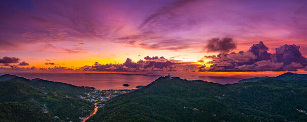 Wall Mural - Aerial hyper lapse view of A breathtaking sunset paints the sky in vivid hues of orange and blue over a coastal town, with lush green hills framing the scene. cloud in dramatic sunset over the ocean