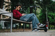 © qunica.com - A young man wearing sunglasses is sitting and relaxing on a park bench, using his phone. An electric scooter is resting beside him, creating a casual and tech-savvy atmosphere.