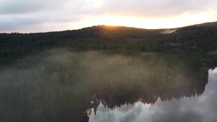 Sticker - Aerial view flying through low clouds over dense forest at sunset, with golden light breaking over the hills
