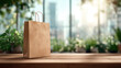 © Denniro - Brown paper bag on wooden table by window. Eco-friendly kraft paper shopping bag with handles placed on a sunlit wooden surface surrounded by green plants and urban window view