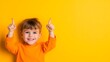 © Koplexs-Stock - Smiling child lying on yellow background pointing upwards with both hands.