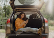 © olezzo - Happy young woman using smartphone sitting in open trunk of car during travel