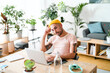 © Studio Marmellata - man with a yellow beanie and tattoos, wearing a striped shirt, works intently on his laptop at a modern wooden desk. He is deep in thought, surrounded by lush green plants in a home office.
