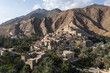 © AmazingAerialAgency - Aerial view of an ancient village nestled at the foot of rugged mountains, a tapestry of stone structures amidst verdant palms, Muscat, Muscat Governorate, Oman.