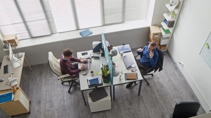 Wall Mural - High angle shot of office employee typing on laptop while male colleague talking on phone call working at shared desk with privacy panel in contemporary workspace