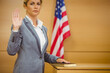 © WavebreakMediaMicro - Woman standing in witness stand raising right hand on Bible beside US flag in grey suit