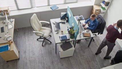 Wall Mural - High angle shot of male manager walking around office room equipped with partitioned desk talking on smartphone then passing phone to colleague during business planning conversation