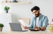 © Prostock-studio - Paying bills, taxes at home online. Young arab man using calculator and laptop computer, calculating taxes or planning budget while sitting at kitchen table at home, copy space