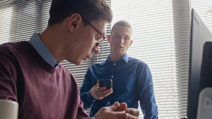 Wall Mural - Focused male office worker with smartphone in hands talking to colleague debating work strategies while collaborating and exchanging ideas in modern workspace