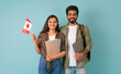 © Prostock-studio - Education abroad, immigration to EU, language school. Cheerful multicultural young arab man and indian woman students with Canadian flag, books and laptop on blue background