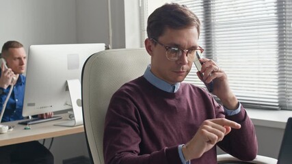 Wall Mural - Medium shot of focused male manager typing on smartphone screen then starting talking on phone call with client while working at desk in office workspace