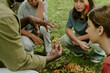 © AnnaStills - Black man teacher showing group of multiethnic children mushrooms outdoors in park, children sitting on grass attentively observing and listening during educational nature lesson