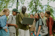 © AnnaStills - Black male teacher showing laptop to group of multiethnic children standing outdoors in park, children watching screen attentively, backpacks visible, trees in background, educational activity