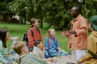© AnnaStills - Group of diverse children sitting on grass listening to Black male teacher explaining lesson outdoors in park, children appearing engaged and attentive, backpacks visible, trees in background