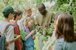 © AnnaStills - Black male teacher showing group of multiethnic children plants in park, children observing and interacting with leaves, outdoor educational activity, learning about nature together