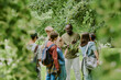 © AnnaStills - Black middle aged man teaching group of multiethnic children outdoors in park, children listening attentively and observing plants, backpacks visible, educational activity taking place