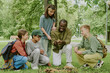 © AnnaStills - Black male young adult teacher showing mushrooms to group of diverse children and teenagers sitting and kneeling on grass in park, students observing and learning outdoors together