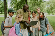 © AnnaStills - Black male teacher showing laptop to group of multiethnic children outdoors in park, children standing closely and watching screen, backpacks visible, learning together during outdoor school activity