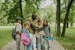 © AnnaStills - Group of diverse children and Black male teacher standing outdoors in park, teacher showing something on laptop, children watching attentively, some holding backpacks and skateboard