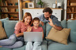 © Stockphotodirectors - A mother, father, and daughter gather around a laptop on a couch, sharing excitement and smiles as they interact with something on the screen, enjoying quality family time.