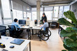 © Koldo_Studio - Businesswoman pointing at computer screen, explaining work to young male employee in wheelchair, working together in modern coworking space