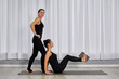 © TRAVELARIUM - Two young adult women in black athletic wear engage in yoga practice. One, seated on mat, elevates her legs with support from block, assisted by other, creating calm, focused atmosphere