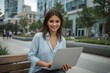 © Paull - Businesswoman in casual wear holding laptop, working while sitting on bench with business center on background.
