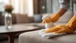 © Prostock-studio - A person cleans the living room furniture with a cloth and spray bottle, wearing yellow gloves.