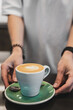 © pavel siamionov - Barista serving cappuccino with latte art in a green saucer on a table. Close-up of hands, spoon, and ceramic cup. Coffee moment in a cozy cafe setting.
