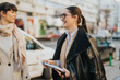 © qunica.com - Two professional women sharing a lighthearted moment outdoors in an urban street setting.