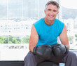© WavebreakMediaMicro - Middle-aged man sitting on bench by glass windows in gym wearing turquoise top and boxing gloves