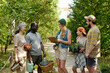 © pressmaster - Group of multiethnic adults and teenager standing outdoors in park, smiling and talking while holding gardening tools, basket, apples, clipboard and potted plants during community activity