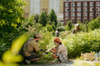 © pressmaster - Caucasian man helping Caucasian girl planting sapling in urban garden while senior Caucasian woman and Black man tending plants in background, group participating in community gardening