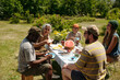 © pressmaster - Group of multiethnic adults and teenagers sitting outdoors around picnic table sharing food and laughing, senior Caucasian woman smiling, Black man holding plate, summer garden setting