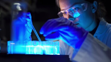 Poster - Female scientist wearing protective glasses and gloves using a micropipette while conducting a research experiment in a dark laboratory with blue light