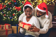 © Prostock-studio - My personal Santa. Happy black woman receiving Christmas gift from her caring boyfriend while sitting near Xmas tree at home