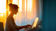 © Digital Design - A woman with glasses works on a laptop at a desk, illuminated by warm sunlight streaming through a window in a calm, cozy room.