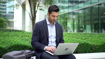 Focused Man in Dark Suit Types on Laptop Outdoor with Building Background