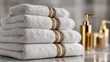 © SSP - A stack of neatly folded white towels with gold trim sits on a reflective surface, accompanied by elegant gold soap dispensers in a soft-focused bathroom setting.