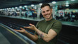 © Krakenimages.com - Young man with tattoos smiling at a busy train station, pointing towards the track with other passengers in the background under bright lights.