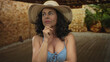 © Krakenimages.com - Middle-aged hispanic woman in a blue bikini with hat indoors appears thoughtful in a rustic setting, emphasizing tranquil summer vibes.