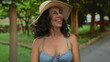 © Krakenimages.com - Woman smiling outdoors in a park wearing a blue bikini and straw hat, middle-aged hispanic enjoying a sunny day amid lush greenery and pathways.