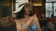 © Krakenimages.com - Hispanic woman in a blue bikini and sun hat gestures indoors at a stylish restaurant setting, conveying her casual yet confident demeanor.