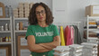 © Krakenimages.com - Hispanic woman volunteer in a green uniform stands confidently indoors in a charity room surrounded by donation items and packaged food.