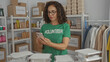 © Krakenimages.com - Middle-aged hispanic woman in volunteer uniform using phone in charity room with donations and supplies neatly organized on shelves.