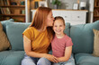 © Stockphotodirectors - A mother kisses her daughter on the forehead while they sit together on a couch, radiating love and joy in their inviting living room filled with books and decorative items.