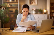© Stockphotodirectors - A woman sits at her home office desk in the afternoon, thoughtfully examining financial documents and bills, showing signs of concern about her financial situation.