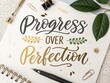 © Mizanur - Inspirational handwritten calligraphy quote progress over perfection on a notebook with pens and leaves