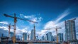© andyaziz6 - Tower cranes dominate the skyline above a bustling construction site as new buildings rise against a vibrant blue sky filled with wispy clouds.